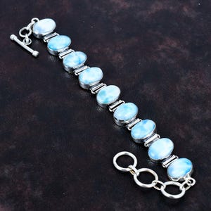 以下が含まれることがあります： 楕円形の水色の宝石をあしらったシルバーブレスレット。 ブレスレットにはトグルクラスプが付いており、暗い表面に表示されています。 宝石はシルバーの金属セッティングにセットされています。