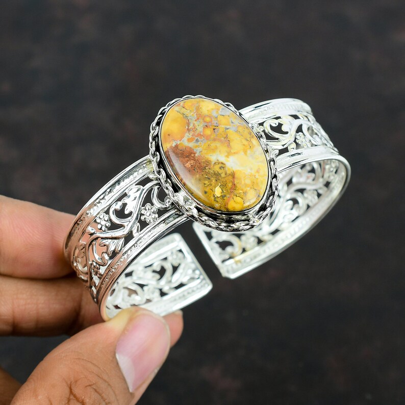 May include: A silver cuff bracelet with an oval-shaped yellow and brown gemstone set in a silver bezel. The bracelet has a delicate filigree design.