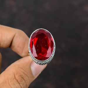 Könnte beinhalten: Ein Silberring mit einem großen, oval geschliffenen, tiefroten Edelstein. Der Edelstein ist der Mittelpunkt, gefasst in einem detaillierten Silberband mit strukturiertem Rand. Der Ring wird vor einem dunkelbraunen Hintergrund gehalten.