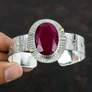 Può includere: Un braccialetto rigido in argento con un grande rubino di forma ovale incastonato in una cornice in argento testurizzato. Il braccialetto ha una finitura martellata e piccoli accenti dorati.