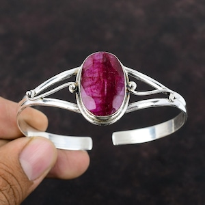 May include: A silver cuff bracelet featuring a large, oval-shaped, deep red gemstone. The gemstone is the focal point, set in a bezel setting. The bracelet has an open design with decorative flourishes on the sides.