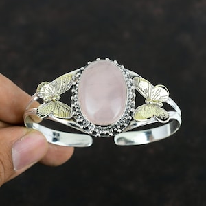 Könnte beinhalten: Silbernes Armreif-Armband mit einem großen ovalen Rosenquarz-Edelstein. Das Armband hat Schmetterlingsakzente mit goldfarbenen Details auf beiden Seiten des Edelsteins. Das offene Design ermöglicht ein einfaches Tragen. Ca. 17 cm Umfang.