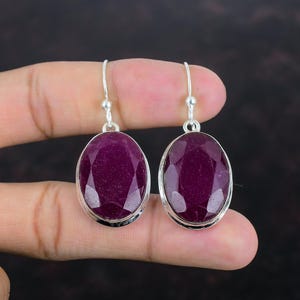 May include: A pair of oval-shaped ruby earrings with a silver setting. The deep red gemstones are faceted, catching the light. The earrings have a simple hook design for pierced ears, showcasing the vibrant color of the rubies.