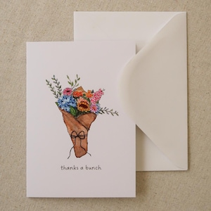 May include: A white greeting card with a watercolor illustration of a bouquet of flowers wrapped in brown paper, tied with a black bow. The card includes the text "thanks a bunch" in a simple font. A white envelope is behind the card.