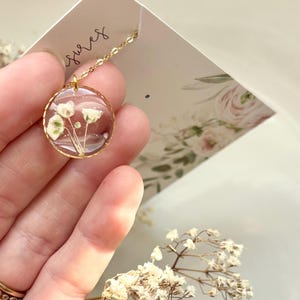 May include: A gold-colored necklace with a round pendant. The pendant contains small white flowers encased in clear resin. The necklace is held by a person. The word "sures" is written on the card in the background.