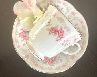 May include: A white teacup and saucer with a floral pattern featuring pink roses and gray leaves. The saucer has a gold trim. A white flower is resting on the saucer.