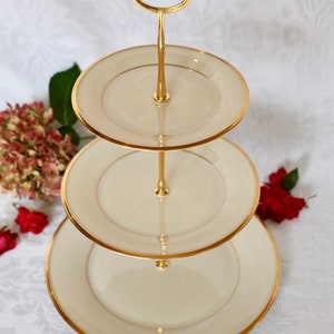 May include: A three-tiered serving stand with a gold metal frame and white porcelain plates with gold trim. The stand has a handle on top for easy carrying.