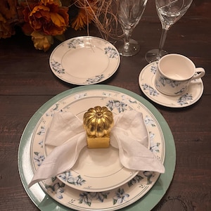 May include: A place setting featuring a white dinner plate and salad plate with blue floral patterns, a white linen napkin held by a gold pumpkin napkin ring, a matching teacup and saucer, two clear stemmed glasses, and a small side plate.