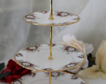 Könnte beinhalten: Ein dreistöckiger Kuchenständer mit einem goldenen Metallrahmen und weißen Porzellantellern. Jeder Teller ist mit einem Blumendesign mit rosa und weißen Rosen verziert.