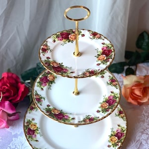 May include: Three-tiered cake stand with a gold metal frame and white porcelain plates decorated with a floral pattern of red, yellow, and pink roses.
