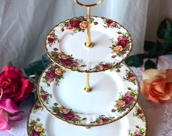 Puede incluir: Un soporte para tartas de tres niveles con un marco de metal dorado y platos de porcelana blanca decorados con un patrón floral de rosas rojas, amarillas y rosas.