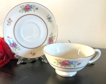 May include: A white teacup and saucer set with floral accents. The cup and saucer have a gold trim and are decorated with pink, blue, and purple flowers. The set is displayed on a black surface.