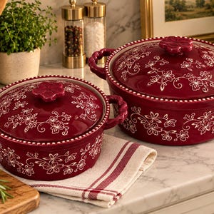 May include: Two round, deep red ceramic cooking pots with matching lids, each featuring a white floral pattern and a beaded rim. The lids have a flower-shaped handle, and the pots have two side handles with a scalloped, rope-like texture.