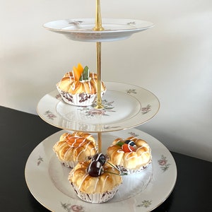 May include: A three-tiered serving stand with floral patterned plates and a gold-colored central support. Each plate holds a small pastry, decorated with fruit. The stand is set against a neutral background, suggesting a display for desserts or appetizers.