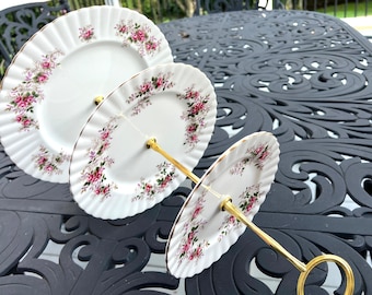 May include: A three-tiered cake stand with a gold metal frame and white porcelain plates with a floral pattern. The plates have a scalloped edge and a gold trim.