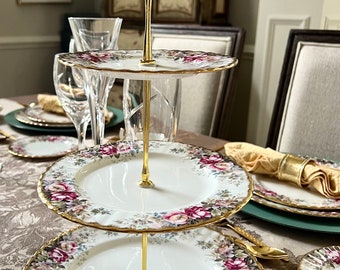 May include: A three-tiered serving tray with a gold handle and support. The white plates are decorated with a floral pattern in shades of pink, purple, and green, with gold trim. The tray is set on a table.