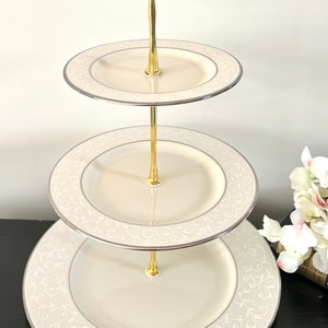 May include: A three-tiered serving stand with a gold-colored handle and support. The plates are cream-colored with a silver trim and a delicate, white floral pattern. The stand is ideal for displaying desserts or appetizers.