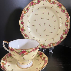 May include: A vintage tea set featuring a teacup, saucer, and plate. The set has a yellow background with a floral pattern and a pink ribbon border. The teacup and saucer have gold trim.
