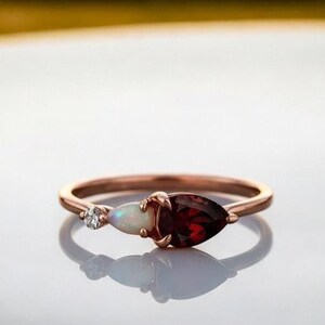 Könnte beinhalten: Ein roségoldener Ring mit einem kleinen Diamanten, einem weißen Opal und einem birnenförmigen, tiefroten Granat. Der Ring ist vor einem verschwommenen Sonnenuntergangshintergrund platziert.