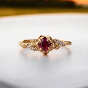 May include: A gold ring featuring a central round red ruby gemstone set within a flower-shaped design. The band is accented with small clear stones and leaf-like metal details extending from the centre setting on both sides of the ring.