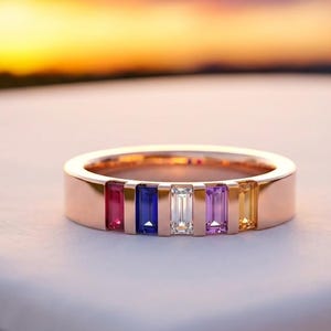Pode incluir: Um anel de ouro rosa com seis pedras retangulares em vermelho, azul, transparente, roxo e amarelo. O anel está sobre um fundo desfocado de um pôr do sol.