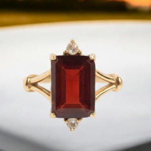 May include: A gold ring with a rectangular, deep red gemstone. Two smaller, clear gemstones are set either side of the main stone. The ring has a classic, elegant design.