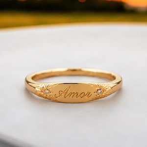 May include: A gold-toned ring featuring a central oval plate engraved with the word Amor in cursive script. The plate is flanked by two small, star-shaped engravings, each containing a tiny embedded clear stone on either side of the text.