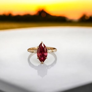 May include: A gold ring with a large, marquise-cut, deep red gemstone as the focal point. The ring's band features small, clear accent stones. The ring is set against a white surface with a blurred sunset background.