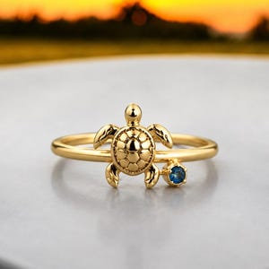 Op de afbeelding: Een goudkleurige ring met een centraal zeeschildpadmotief met een textuur op het schild. Naast de schildpad bevindt zich een enkele, kleine, ronde, diepblauwe edelsteen gezet in een goudkleurige zetting, wat een verfijnd sieraad vormt.