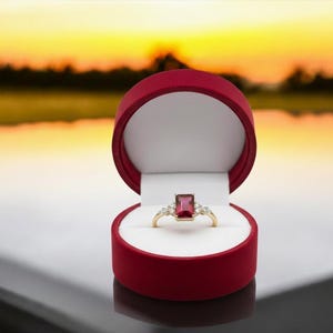May include: A gold ring with a rectangular, red gemstone and smaller white stones, displayed in an open, red ring box with a white interior. The background shows a blurred sunset.