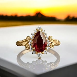 Pode incluir: Um anel de ouro com uma pedra preciosa vermelha escura em forma de pêra, rodeada por pequenas pedras claras. O anel tem um design ornamental e filigranado na banda e em torno da pedra central, contra um fundo desfocado de pôr do sol.