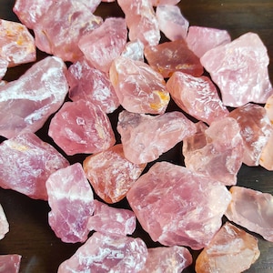 Può includere: Una collezione di cristalli di quarzo rosa, alcuni con un aspetto bianco e nuvoloso. I cristalli sono grezzi e non levigati, con forme e dimensioni irregolari.