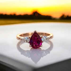 Può includere: Un anello in oro rosa con una pietra preziosa rosso scuro a forma di pera come elemento centrale, affiancata da due pietre preziose chiare più piccole. L'anello è incastonato su uno sfondo sfocato di tramonto.