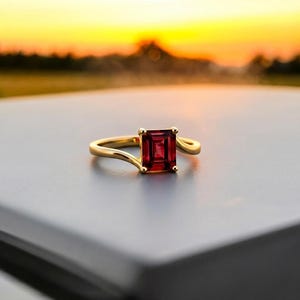 Puede incluir: Un anillo de oro con una piedra preciosa de color rojo intenso en talla esmeralda, sujeta por cuatro garras. La banda tiene un acabado liso y pulido con un diseño ligeramente curvado que se estrecha hacia la piedra central, de aspecto limpio.