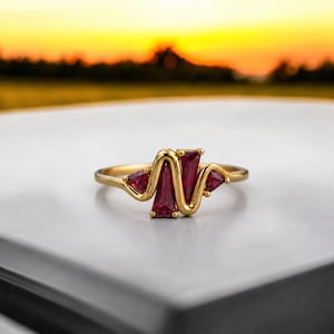 Puede incluir: Un anillo de oro con un diseño de banda ondulada similar a un latido del corazón, engastado con cuatro piedras preciosas de color rojo intenso y corte triangular. La banda de metal tiene un acabado pulido y las piedras están dispuestas.