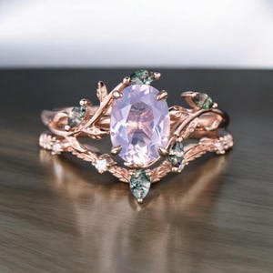 Pode incluir: Um anel de ouro rosa com uma grande gema rosa ovalada cercada por gemas verdes menores e uma banda delicada de diamantes.