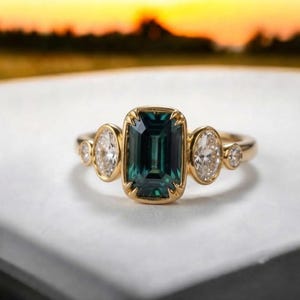 May include: A gold ring featuring a central emerald-cut dark teal gemstone held by prongs. The center stone is flanked on each side by one oval-cut clear gemstone and one smaller round-cut clear gemstone, all set in a polished gold band.