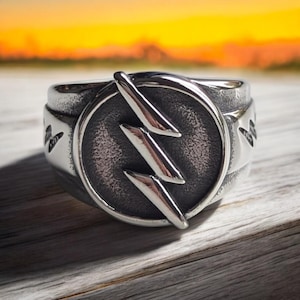 Könnte beinhalten: Ein silberner Ring mit einem schwarzen Blitzsymbol in einem Kreis. Der Ring hat eine strukturierte Band.
