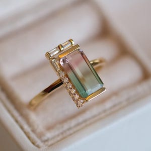 Puede incluir: Un anillo de oro con una piedra preciosa rectangular verde, rosa y azul rodeada de pequeños diamantes. El anillo está engastado en una banda de oro.