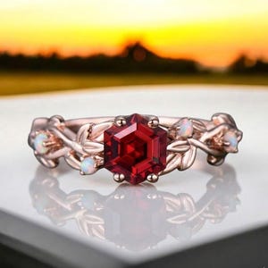 May include: Rose gold ring with a large, faceted, red gemstone at the centre. The ring has a floral design with small, white gemstones and leaf accents. The ring is set against a blurred background.