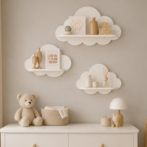 Peut inclure: Trois étagères murales blanches en forme de nuage portant de petits objets décoratifs, dont un panneau indiquant Twinkle Little Star et un autre I Love You To The Moon And Back, au-dessus d'une commode blanche à six tiroirs avec des boutons en bois.