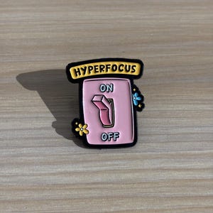 May include: A pink and black enamel pin in the shape of a light switch. The pin has the words "HYPERFOCUS" above the switch, with "ON" and "OFF" on the switch. Small flower accents are on the sides.
