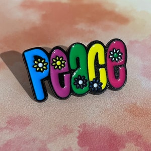 May include: A colourful enamel pin with the word "Peace" spelled out in a groovy font. Each letter is outlined in black and decorated with small, colourful flowers.