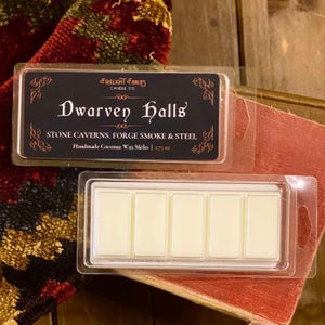 May include: Two clear plastic containers of wax melts. One container is open, revealing five rectangular wax melt pieces. The other container is closed, displaying the text "Dwarven Halls" and "STONE CAVERNS, FORGE SMOKE & STEEL" on a black label. The label also says "Handmade Coconut Wax Melts | 1.75 oz."