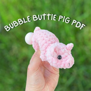 May include: A pink crocheted pig plushie with black eyes. The pig is being held in a hand with a green background.