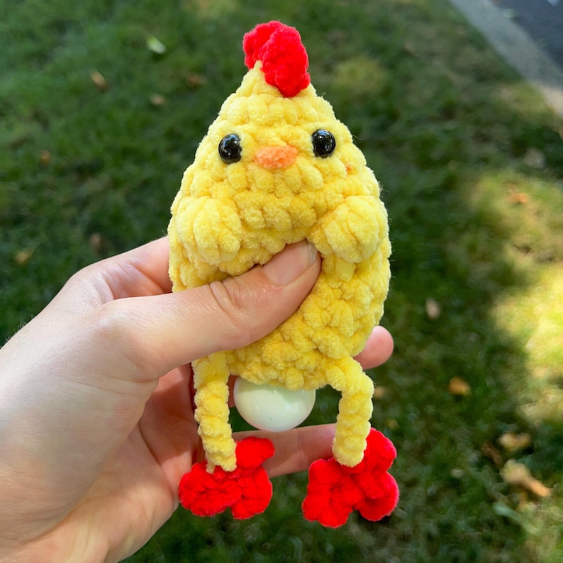 May include: A yellow crocheted chick toy with red feet and a red comb. The chick is holding a white egg in its belly.