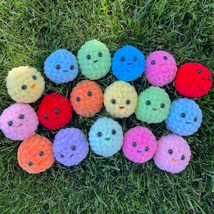 May include: A collection of 17 colourful crocheted plush toys with various facial expressions. The toys are round and have a soft, fuzzy texture. They are arranged in a circle on a green grassy surface.