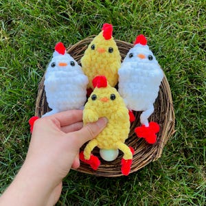 May include: Four crocheted chicks in a wicker nest. Two chicks are white with red combs and feet. Two chicks are yellow with red combs and feet. One yellow chick is holding a white egg.