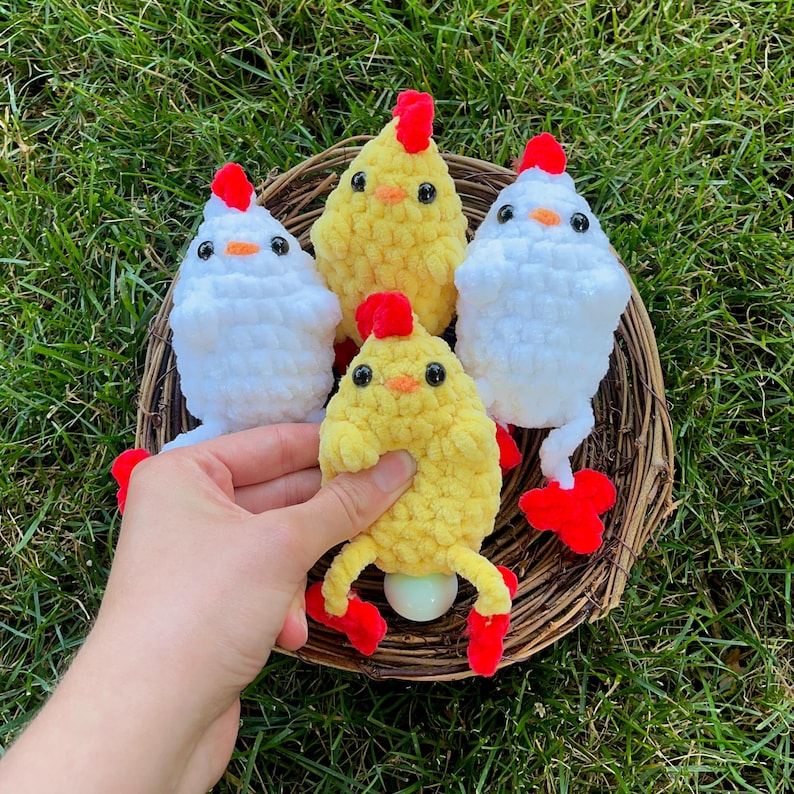 May include: Four crocheted chicks in a wicker nest. Two chicks are white with red combs and feet. Two chicks are yellow with red combs and feet. One yellow chick is holding a white egg.