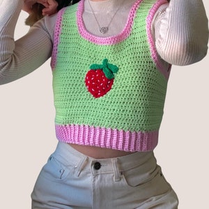 Peut inclure: Un gilet en crochet vert avec une bordure rose et une applique en forme de fraise. Le gilet est sans manches et a un col rond.
